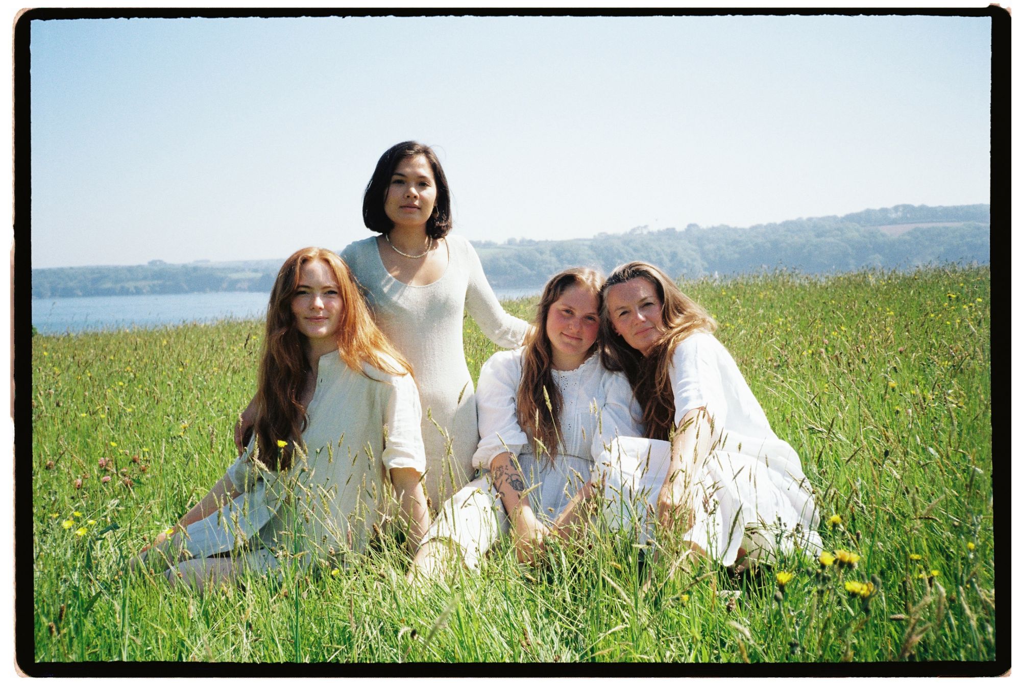 Four women sitting together in long grass above the water, shot on film